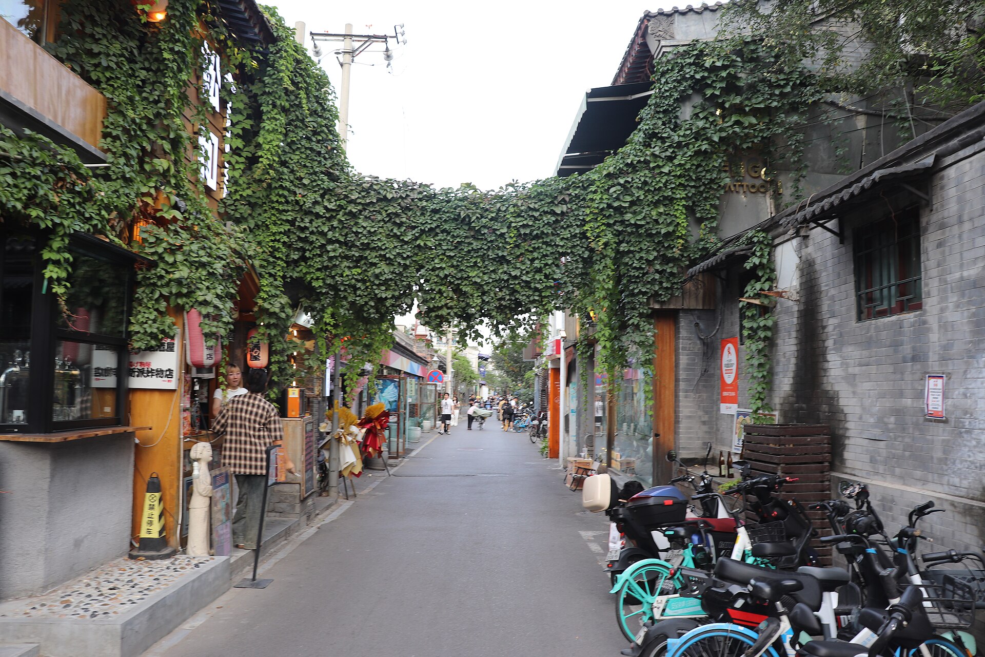 A narrow lane in Beijing's Wudaoying Hutong with grey-brick walls, shops, greenery and parked bicycles in 2023.