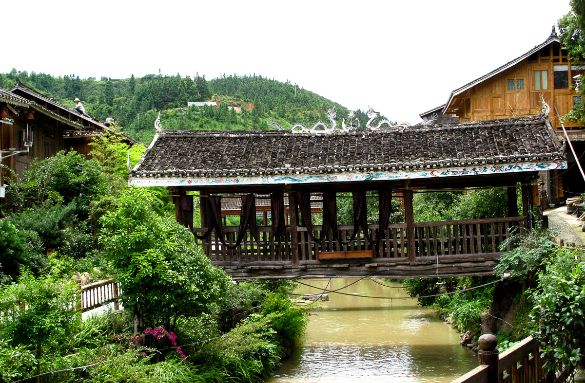 A roofed wooden wind-and-rain bridge crosses a stream between buildings in Zhaoxing.