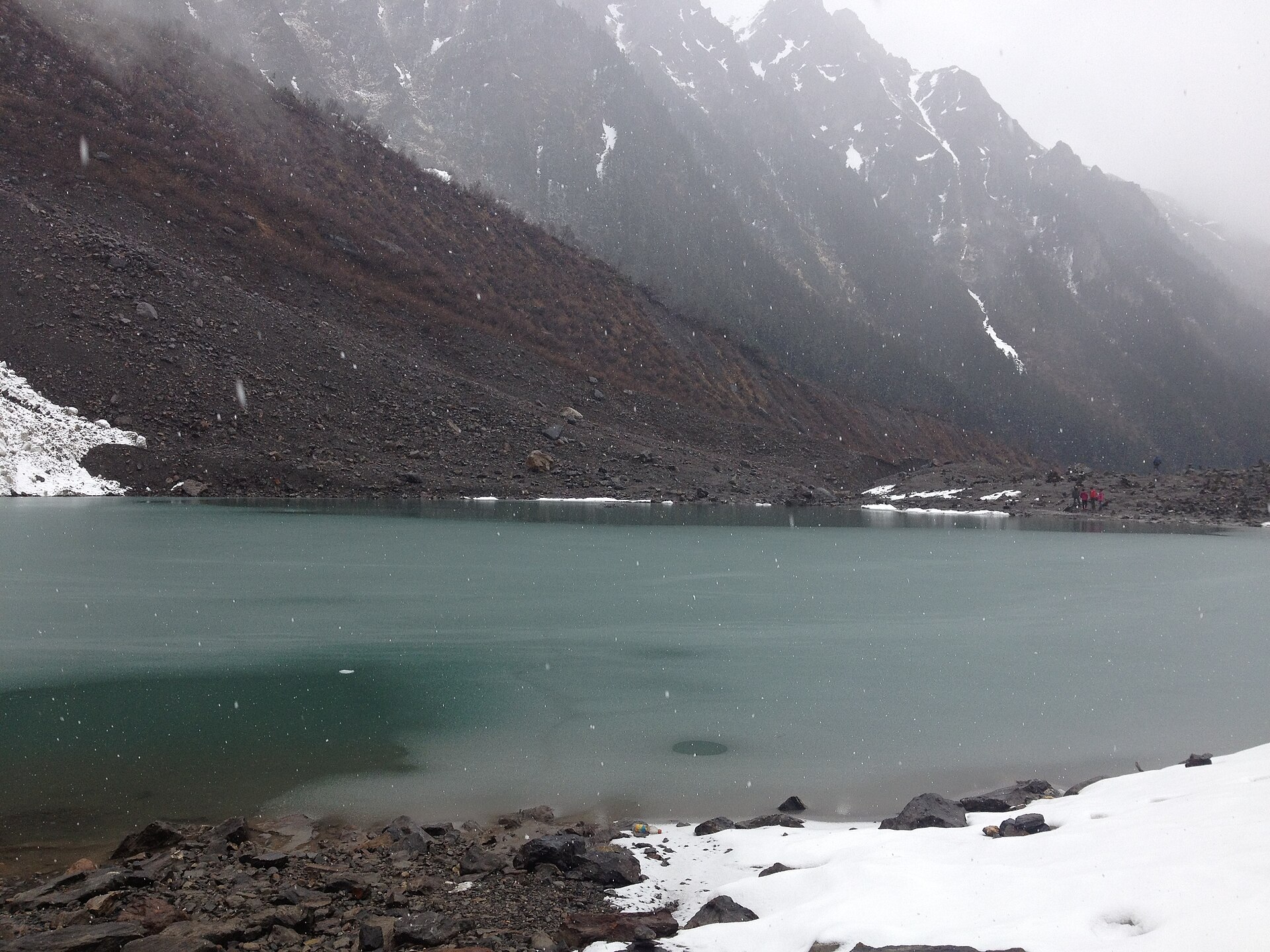 Yubeng Ice Lake during snowfall, with snow on the rocky shore and hikers visible across the water.
